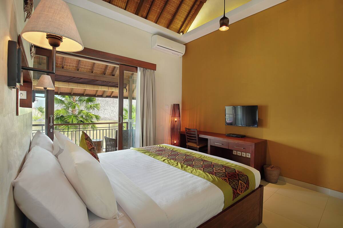 A hotel room with a large bed, a desk, a television, and a balcony with a view of palm trees.