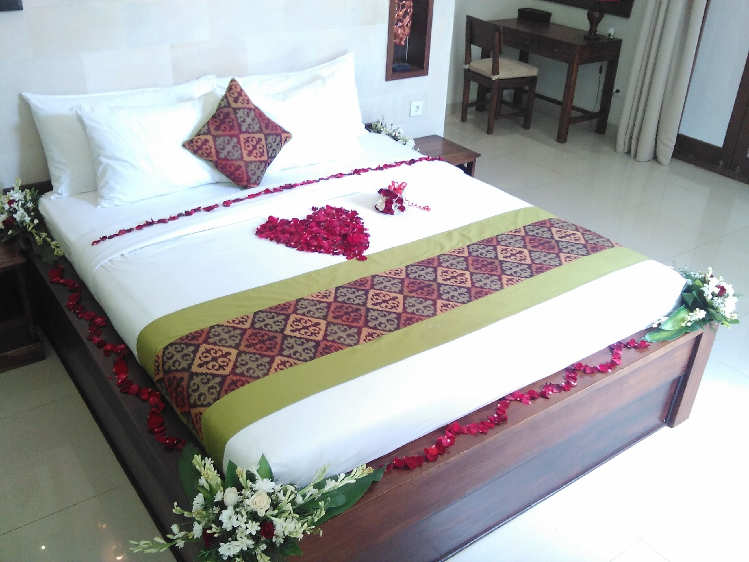 A neatly made bed with a patterned bedspread, a decorative pillow, and a heart-shaped arrangement of red flowers on the bed.