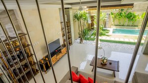 A modern living room with a sofa, coffee table, and TV, overlooking a pool and garden.