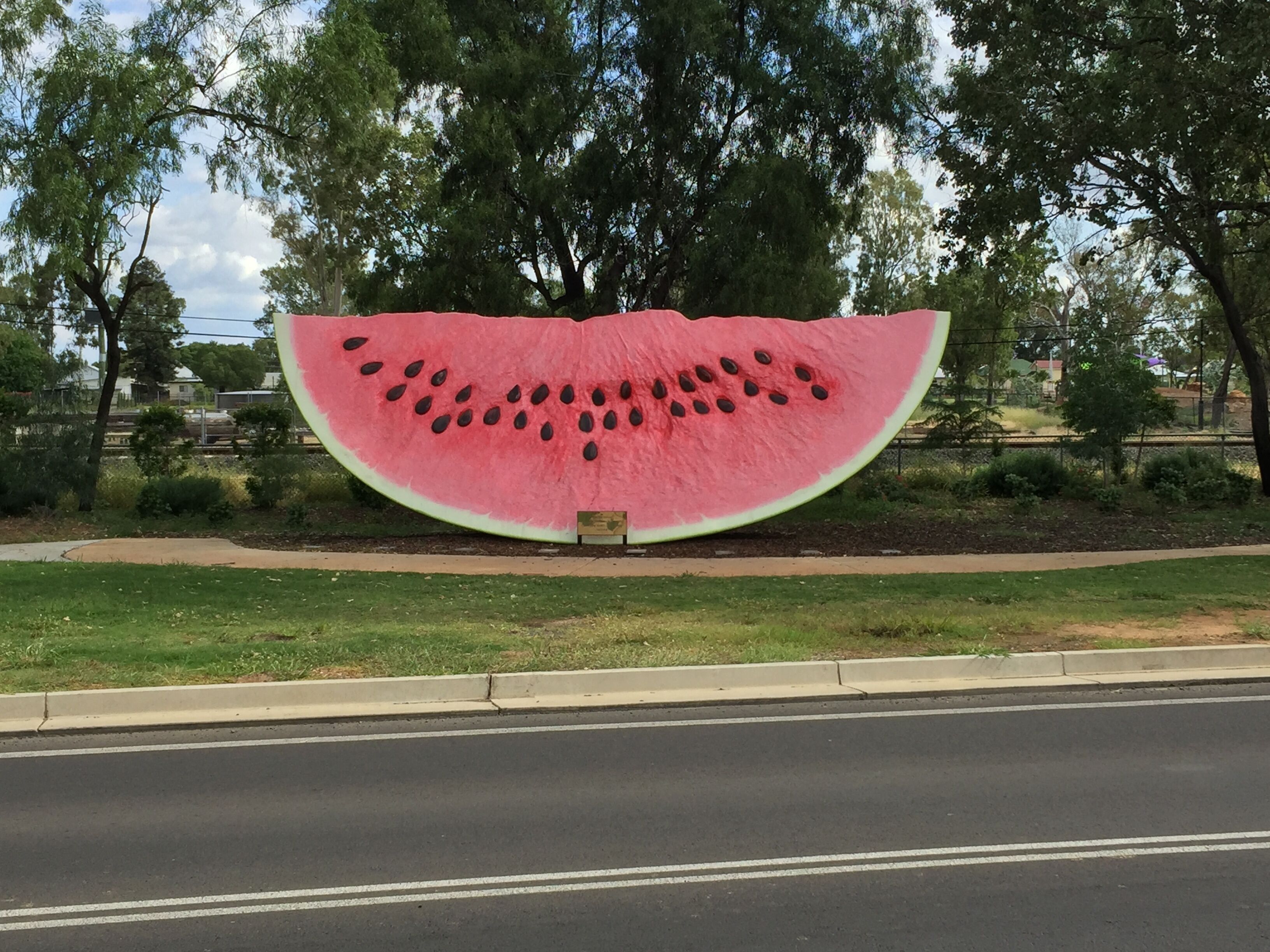 A large watermelon sculpture with seeds, located beside a road with a sidewalk and trees.