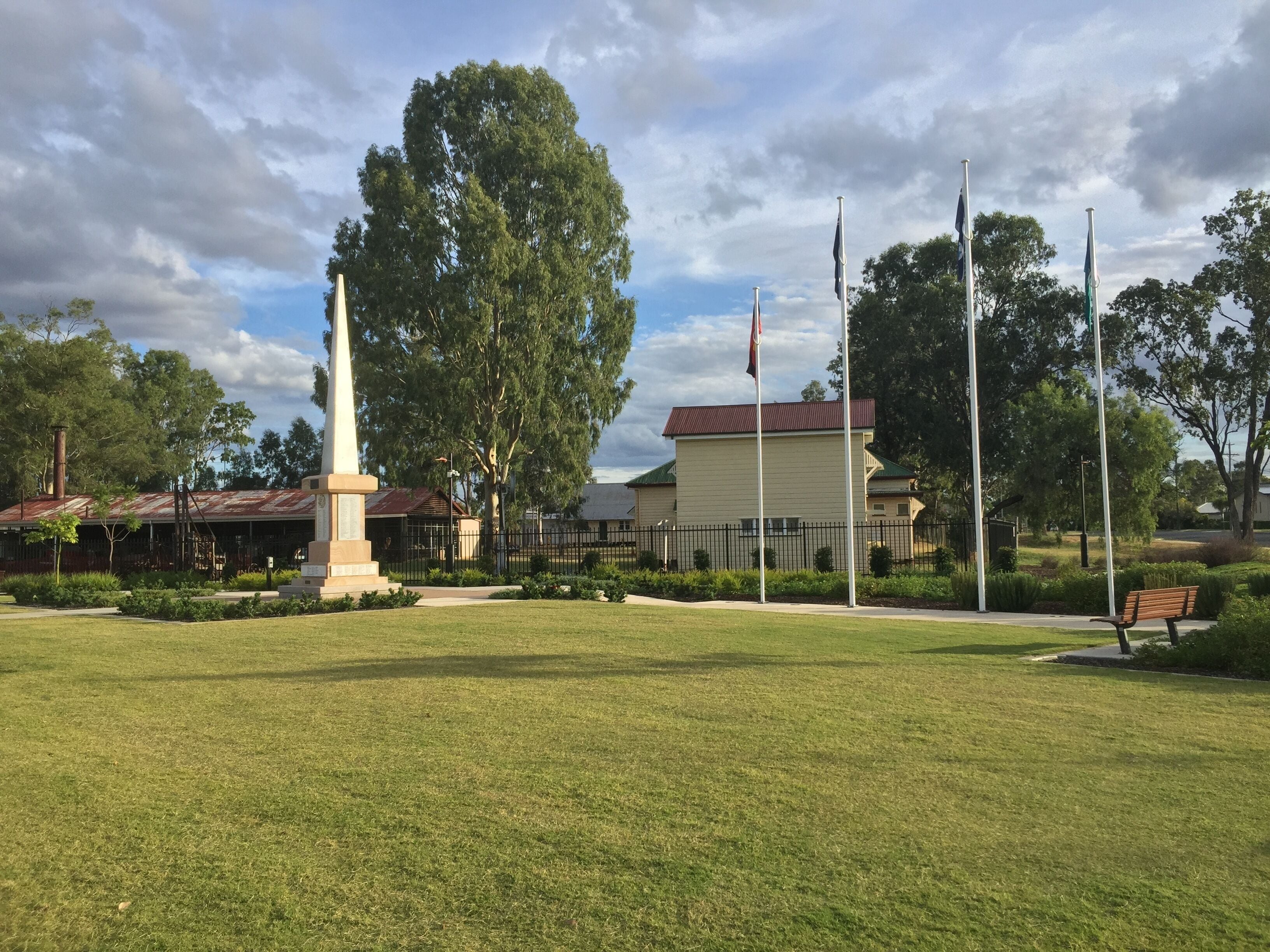 A grassy park with a monument, a bench, and a building with a red roof.