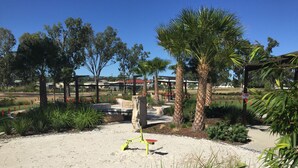 Un parque con zona de juegos de concreto, palmeras y un cielo azul despejado.