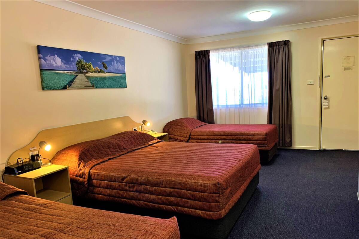 A hotel room with two beds, a painting of a beach scene, a desk with a lamp, and a door with a sign.