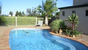 Una piscina con solárium de ladrillo, rodeada por una valla y algo de vegetación.