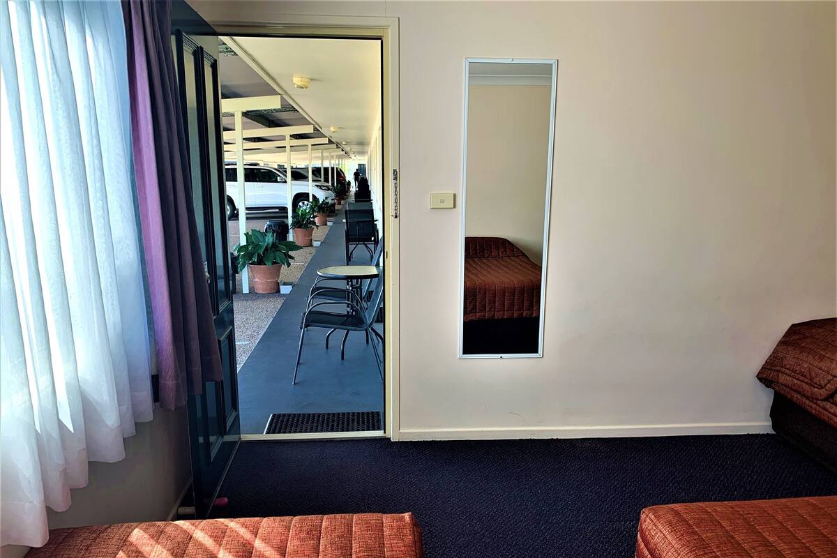 A hotel room with two beds, a small table, and chairs. There is a door leading to a balcony with potted plants and a white car parked outside.