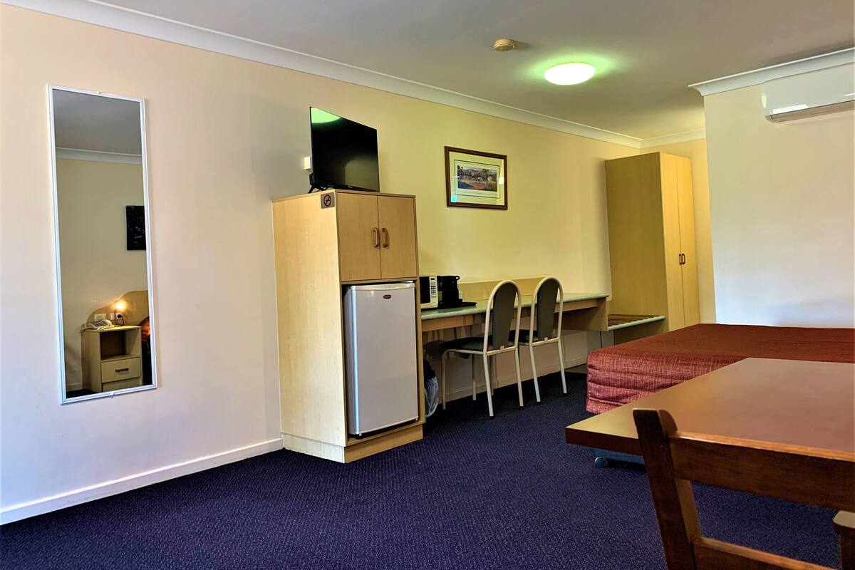 A compact room with a desk, chairs, a small refrigerator, a microwave, and a bed with a red blanket.