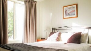 Un dormitorio con cama, cortinas, una mesita de noche y un cuadro en la pared.