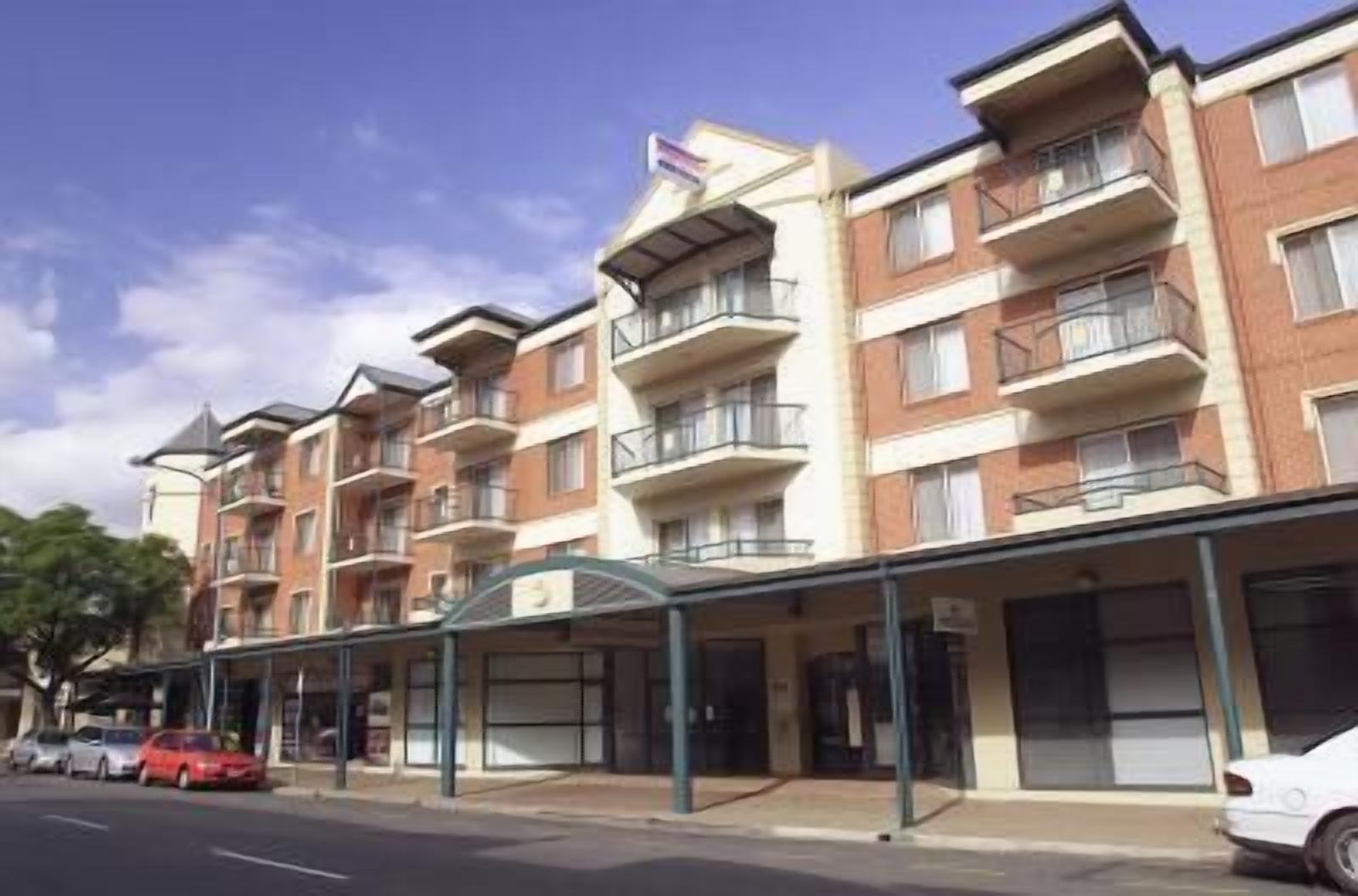 A multi-story residential building with balconies, a covered entrance, and a parking area with cars.