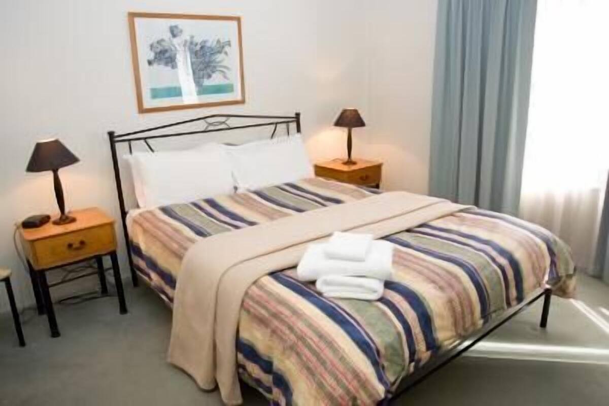 A neatly made bed with striped bedding, two bedside tables with lamps, a framed picture on the wall, and a window with curtains.