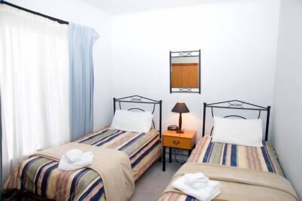 A small, clean room with two single beds, a nightstand with a lamp, and a window with curtains.