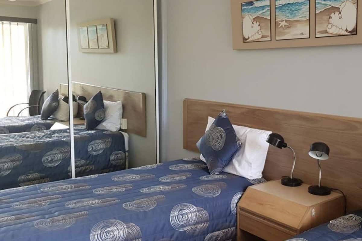 A hotel room with two beds, each with blue bedding and patterned pillows, a wooden headboard, a bedside table with a lamp, and a mirror on the wall.