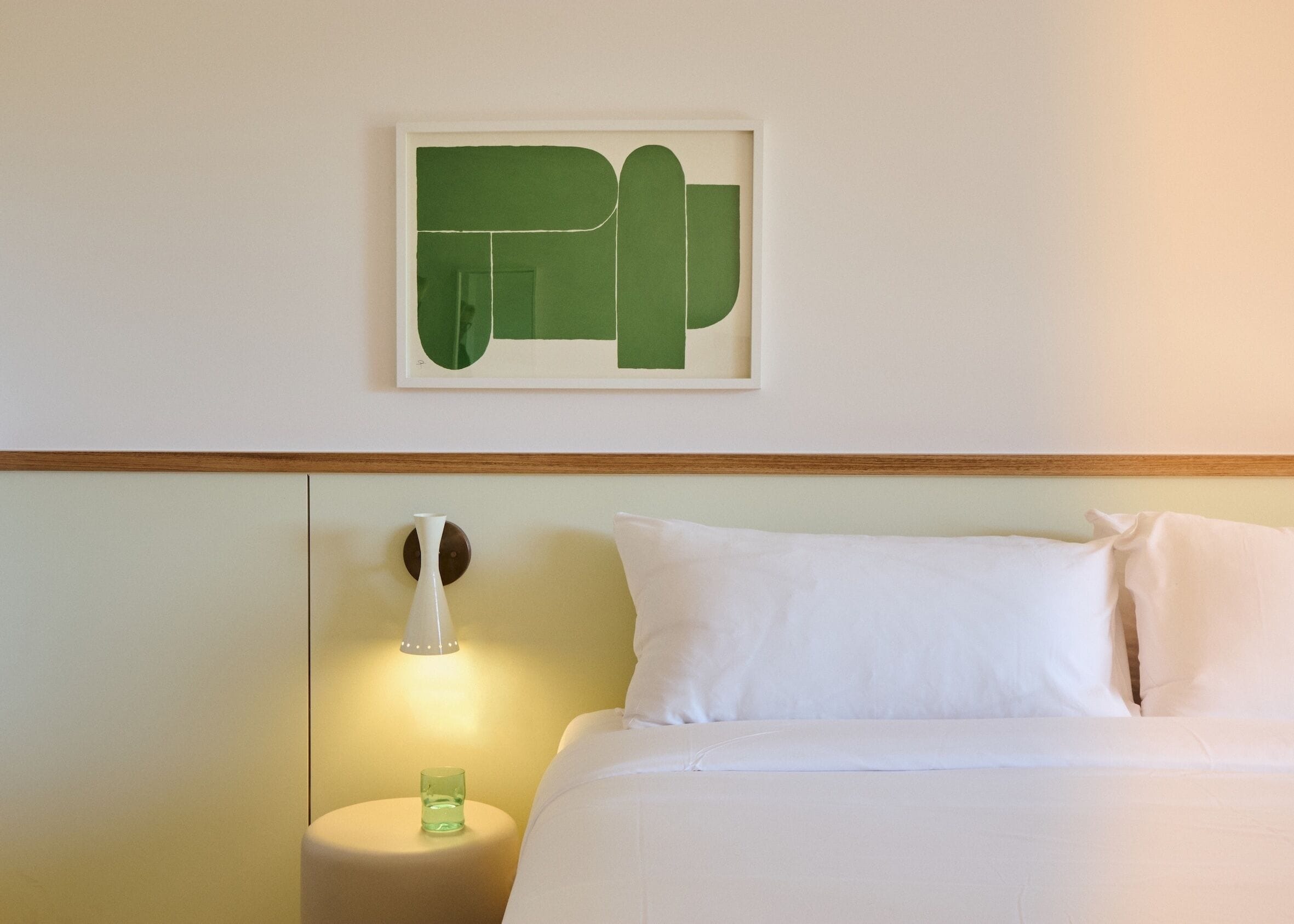 A hotel room with a bed, a nightstand, a wall-mounted lamp, and a framed abstract artwork.