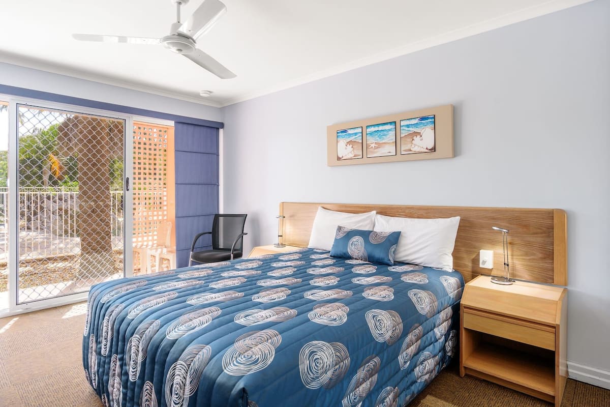 A bedroom with a bed, a nightstand, a chair, a ceiling fan, and a view of the outside.