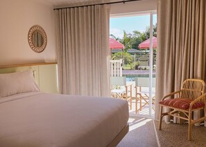 Habitación de hotel con cama, una silla, ventana con cortinas y vistas a un espacio exterior con sombrillas.