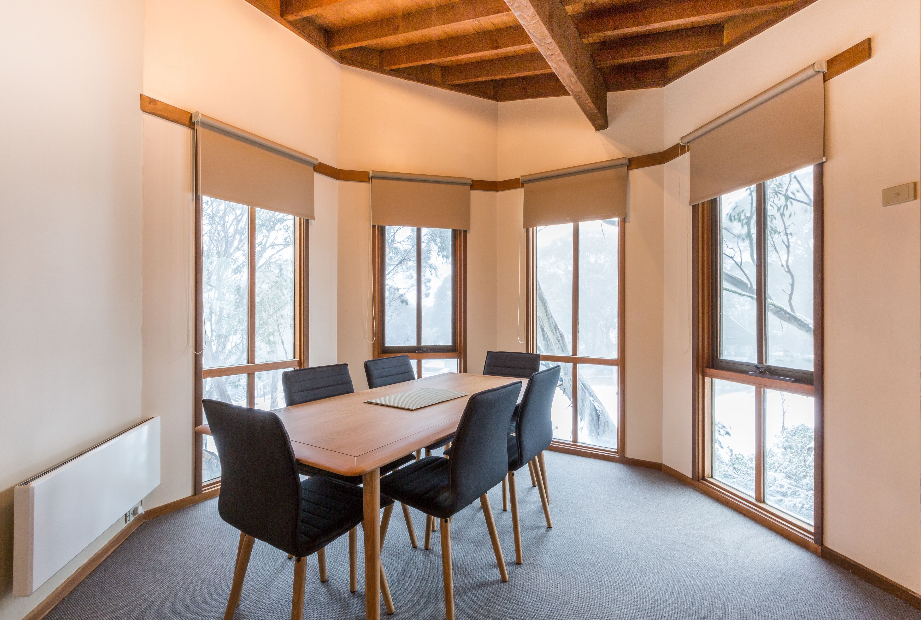 Une salle de conférence avec une table en bois, des chaises noires et de grandes fenêtres équipées de stores.