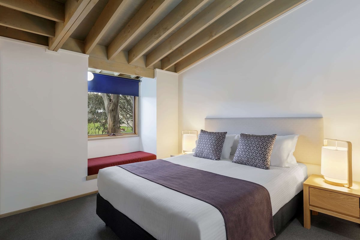 A modern hotel room with a large bed, bedside lamps, a bench, and a window with a view of trees.