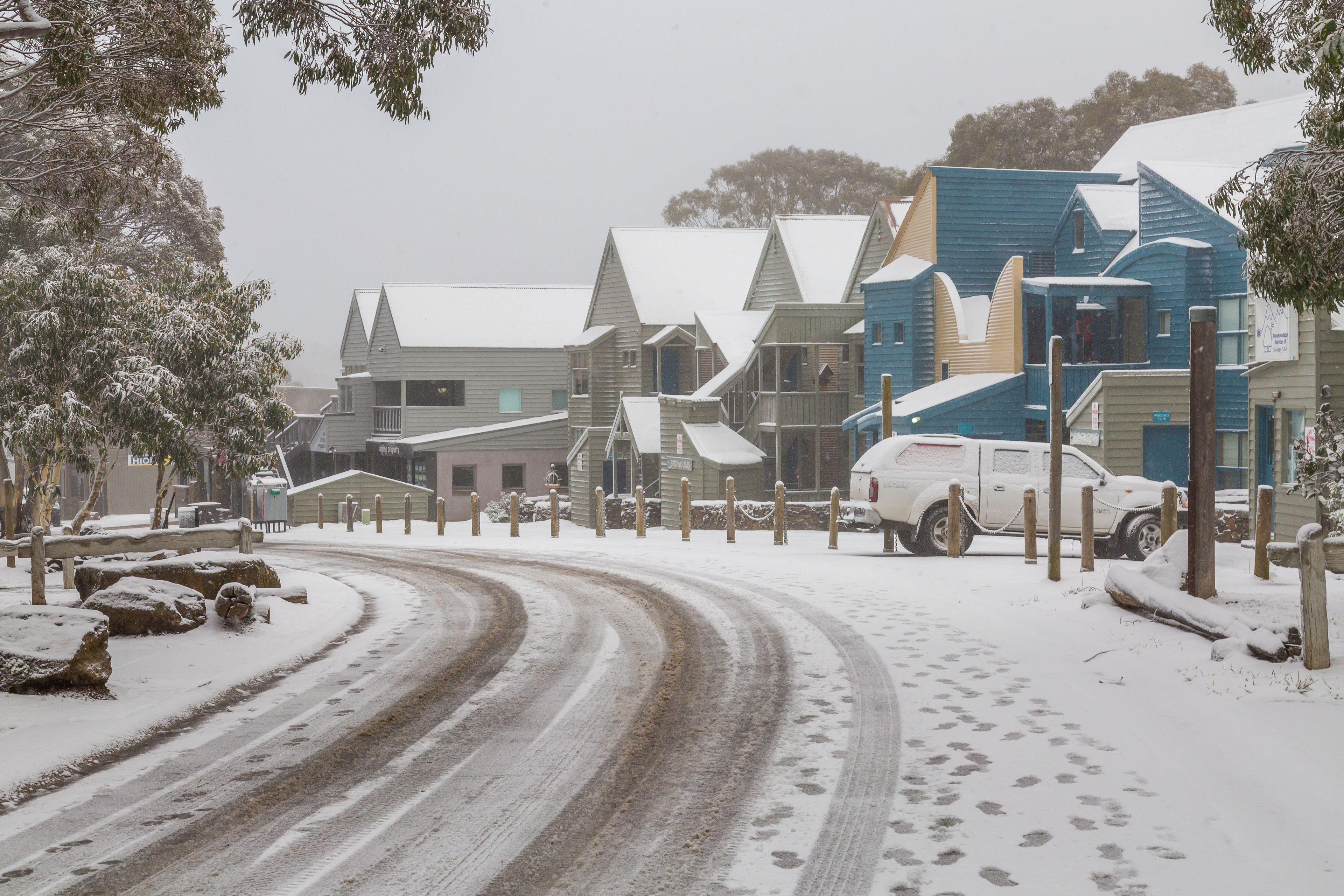 A snow-covered street with houses and a white vehicle parked on the side.