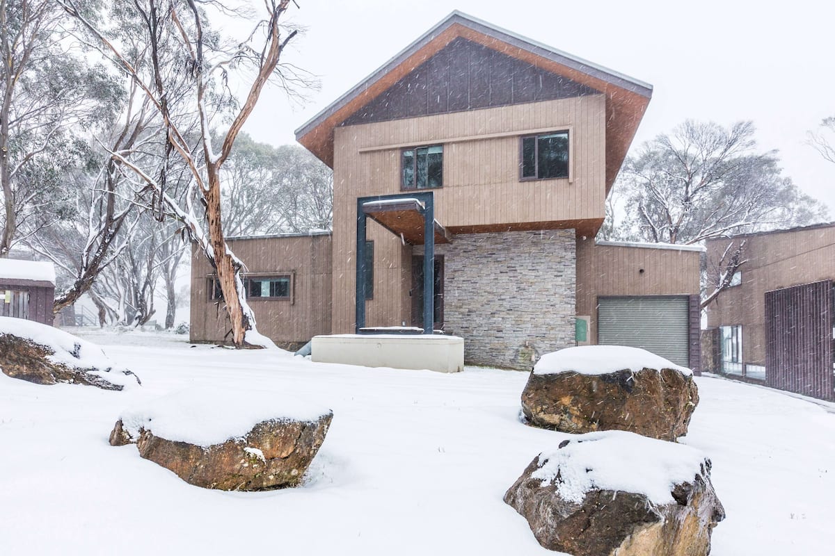 A modern two-story house with a stone facade and wooden exterior, surrounded by snow-covered rocks and trees.