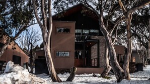 A modern two-story house with a stone facade and large windows, surrounded by snow and trees.