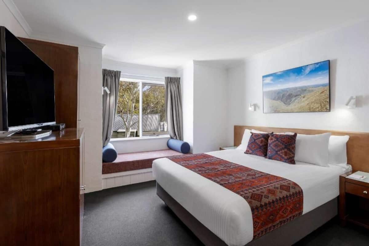 A hotel room with a bed, a TV, a window with curtains, a painting on the wall, and a desk.