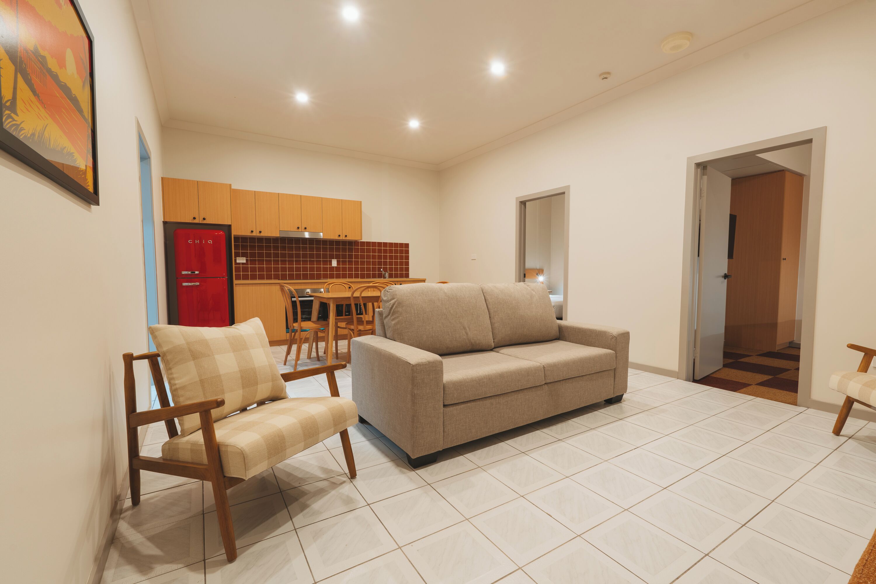 A living room with a sofa, two armchairs, a dining table, and a kitchen area with cabinets and a refrigerator.