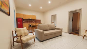 A living room with a sofa, two armchairs, a dining table, and a kitchen area with cabinets and a refrigerator.