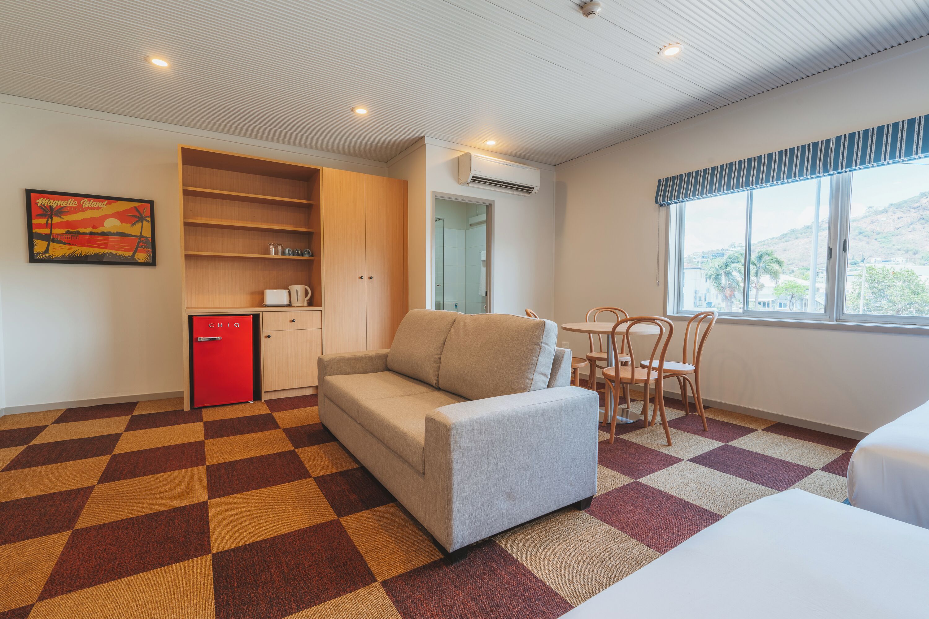 A hotel room with a sofa, a dining table with chairs, a kitchenette, and a window with a view.
