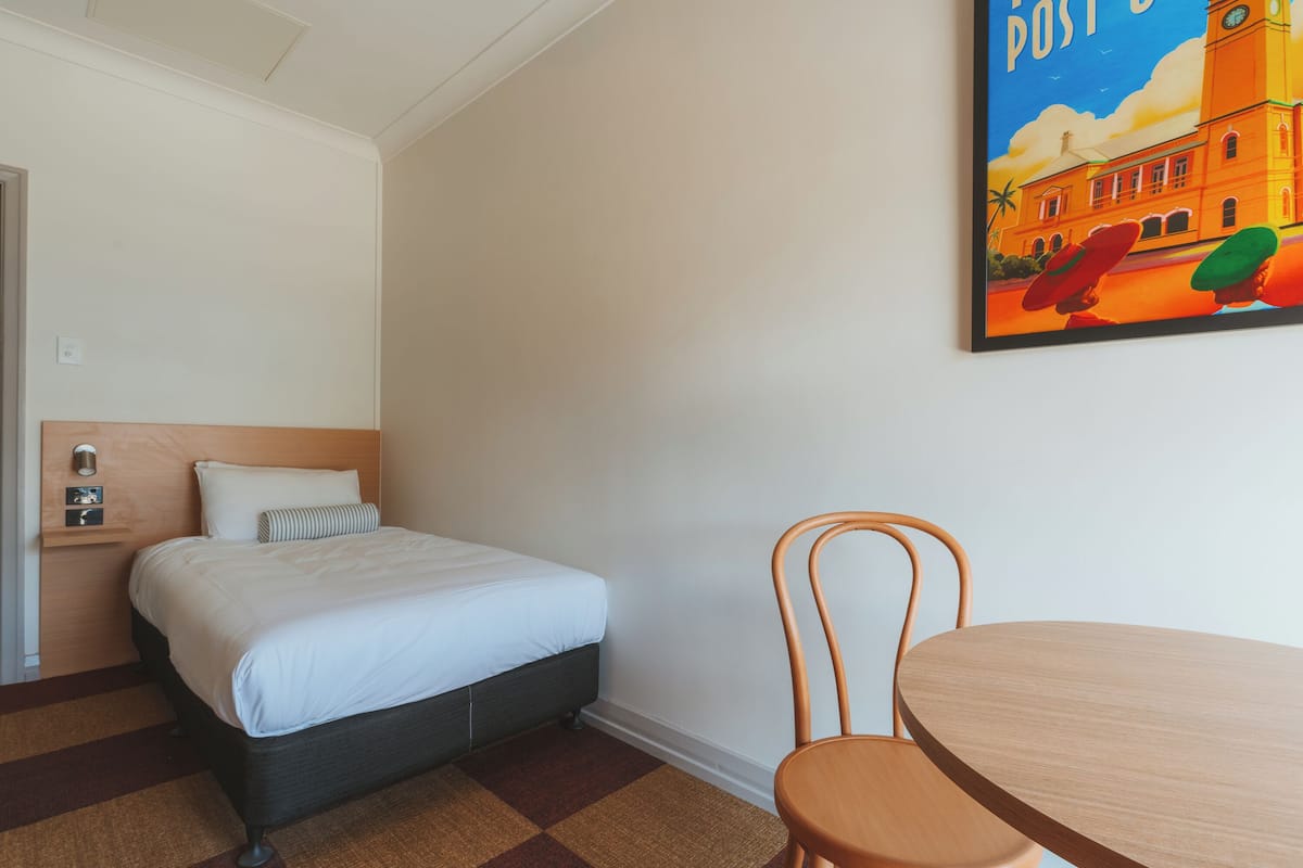 A hotel room with a bed, a chair, a table, and a painting on the wall.