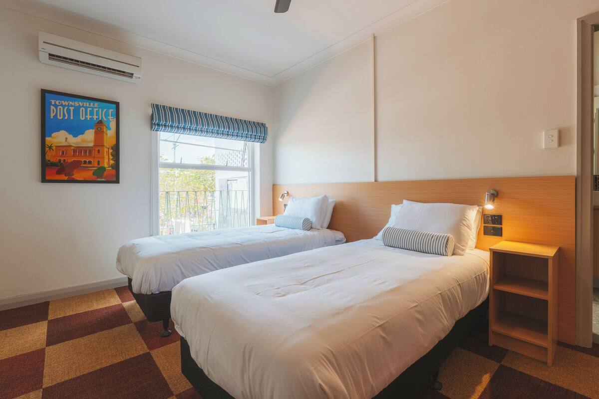 A hotel room with two beds, a window with a striped valance, an air conditioning unit, and a poster of the Townsville Post Office.
