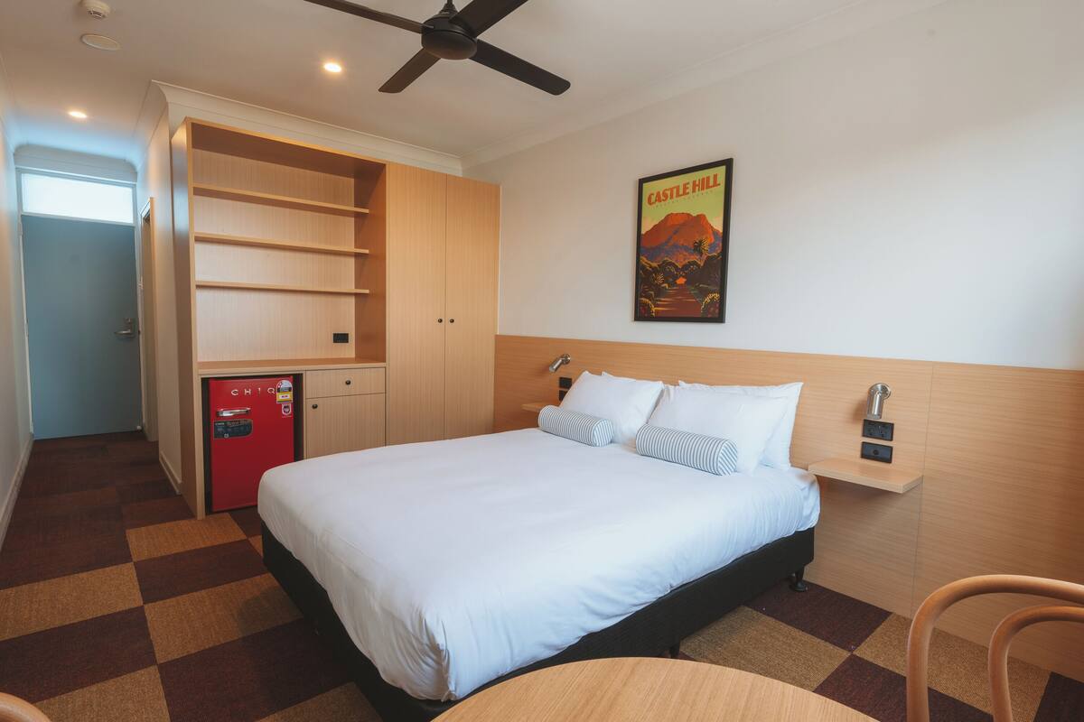 A hotel room with a bed, a small refrigerator, a ceiling fan, a poster on the wall, and a check-in counter.