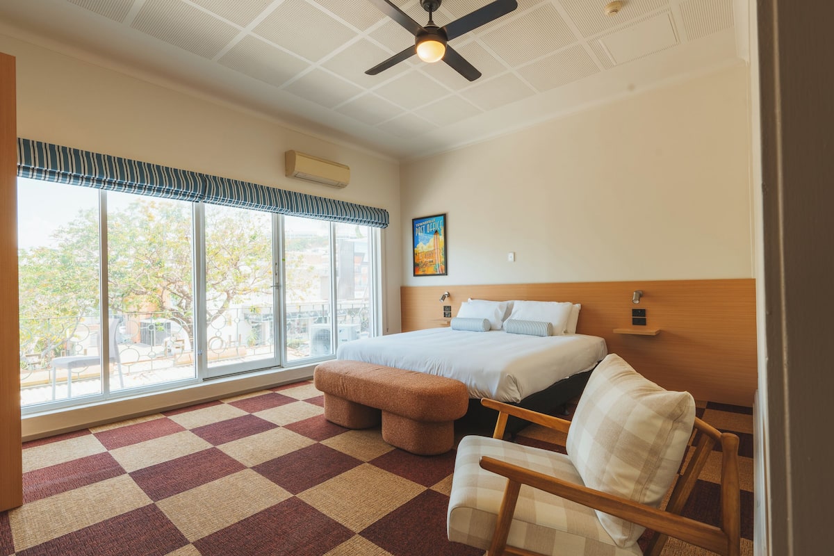 A hotel room with a large window, a bed, a chair, a bench, and a ceiling fan.