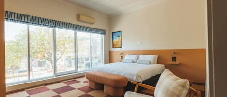 A hotel room with a large window, a bed, a chair, a bench, and a ceiling fan.