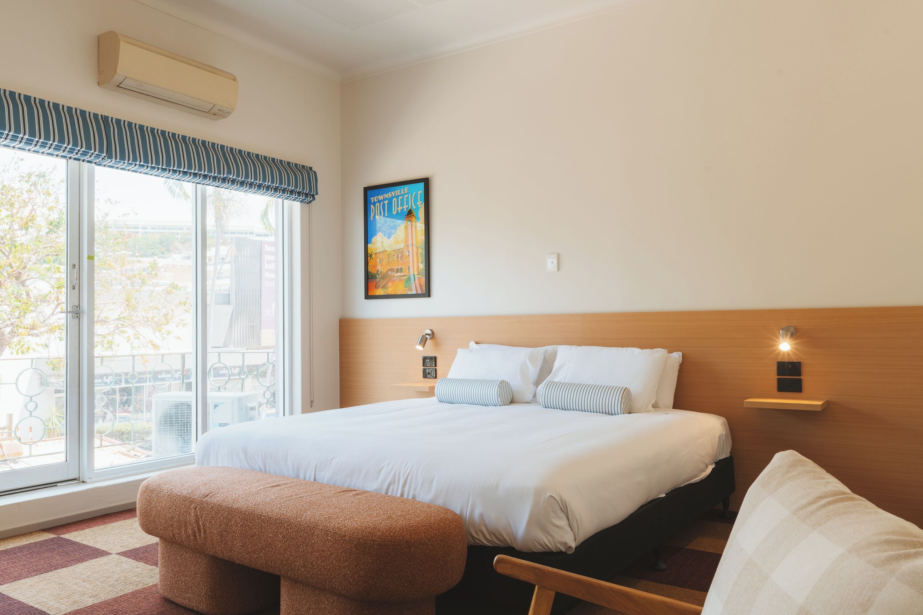 A hotel room with a large bed, a bench, a chair, a window with a view, and a wall-mounted air conditioner.