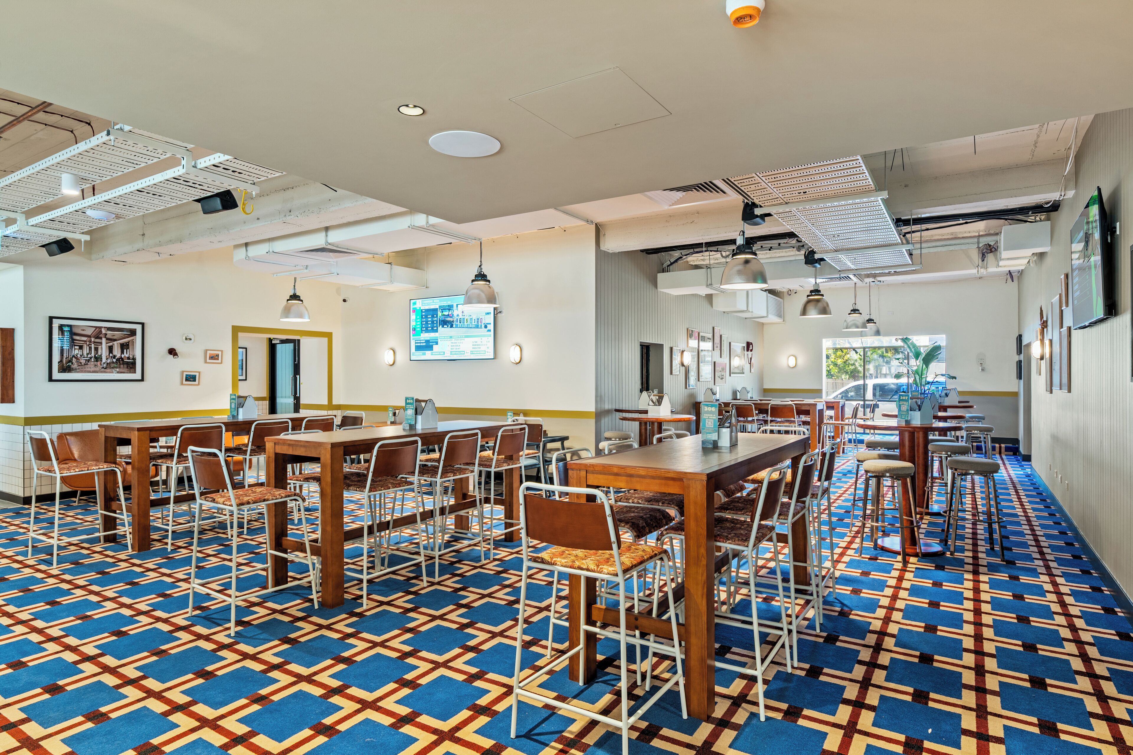 A spacious dining area with multiple tables and chairs, a patterned carpet, and a large screen displaying information.