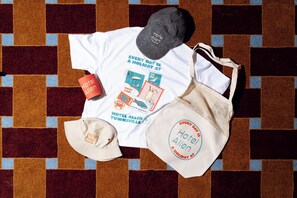 Una camiseta blanca con un estampado de una piscina y una playa, una gorra color beige, una taza roja y un bolso de tela color beige con la inscripción 'Hotel Allen'.