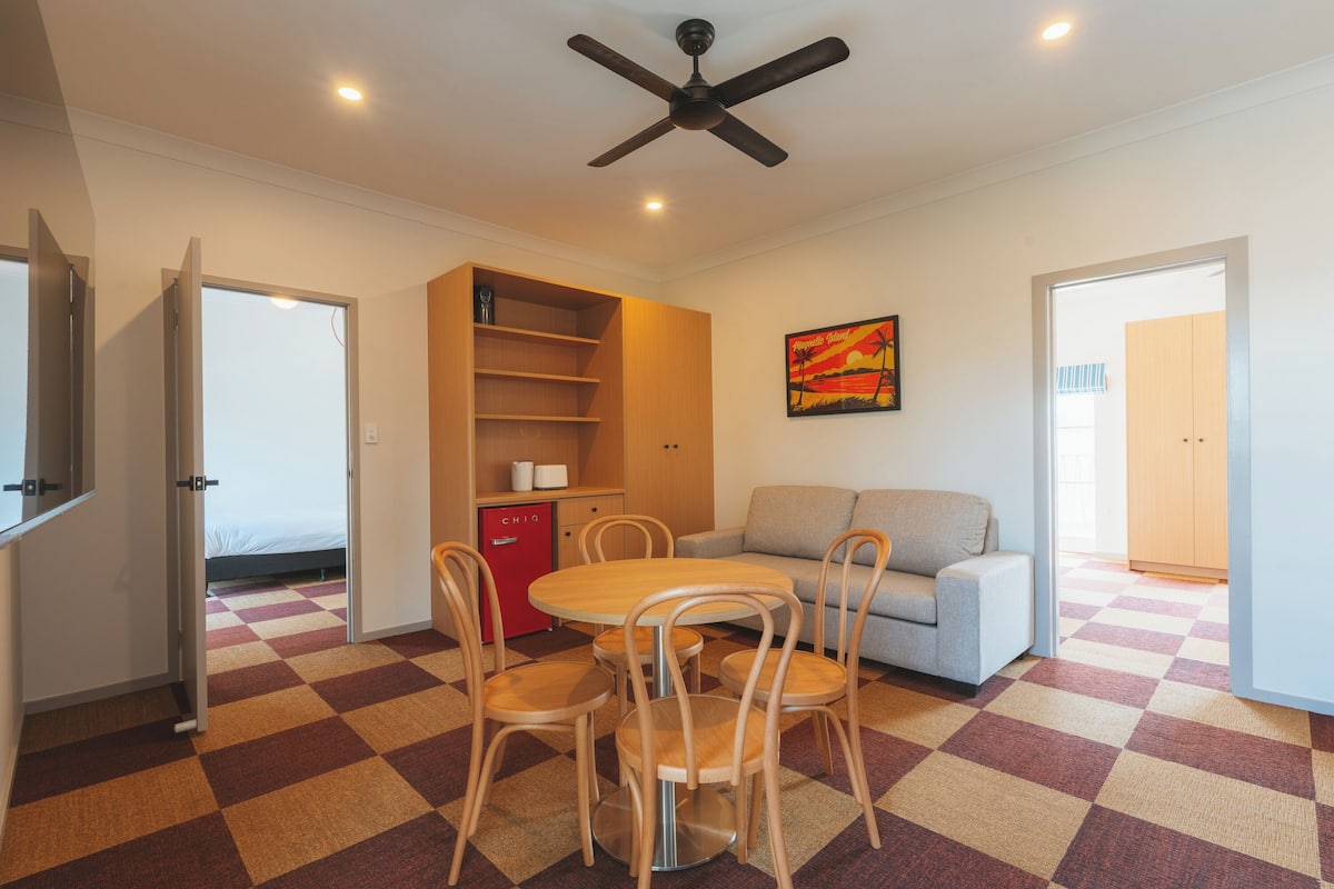 A hotel room with a checkered floor, a dining table with chairs, a sofa, a red minibar, and a ceiling fan.