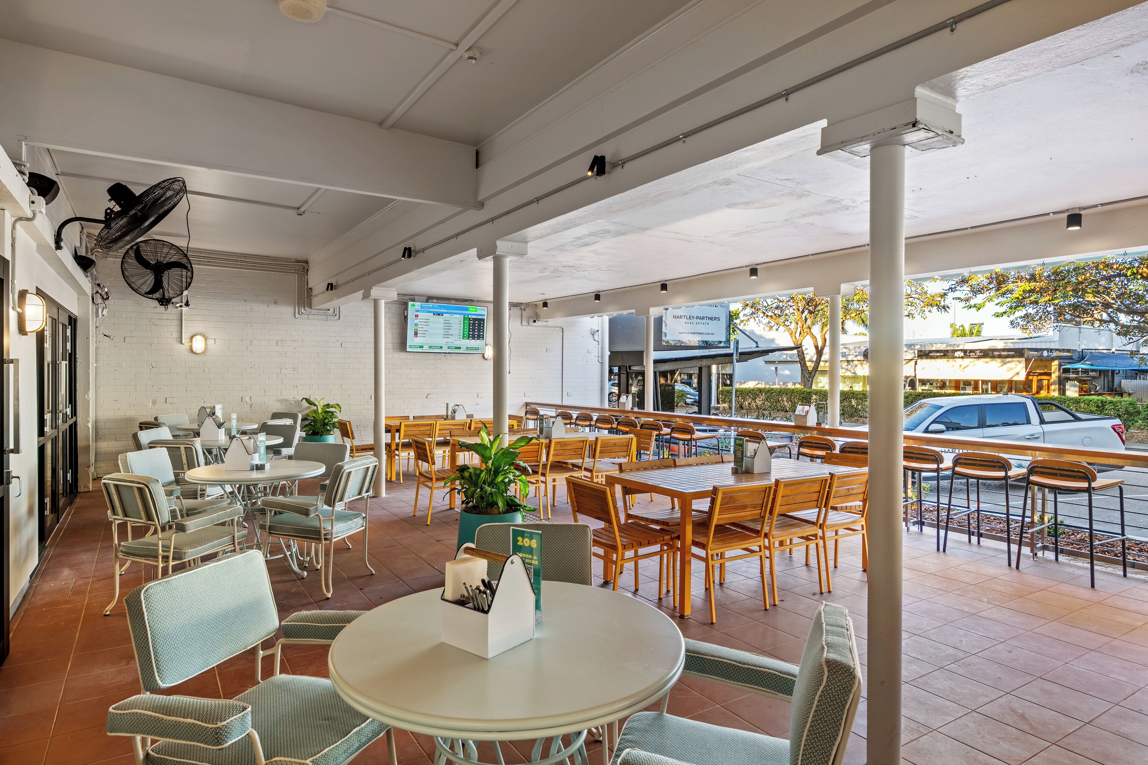 An outdoor cafe with tables and chairs, a TV screen, and a brick wall.