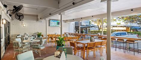 An outdoor cafe with tables and chairs, a TV screen, and a brick wall.