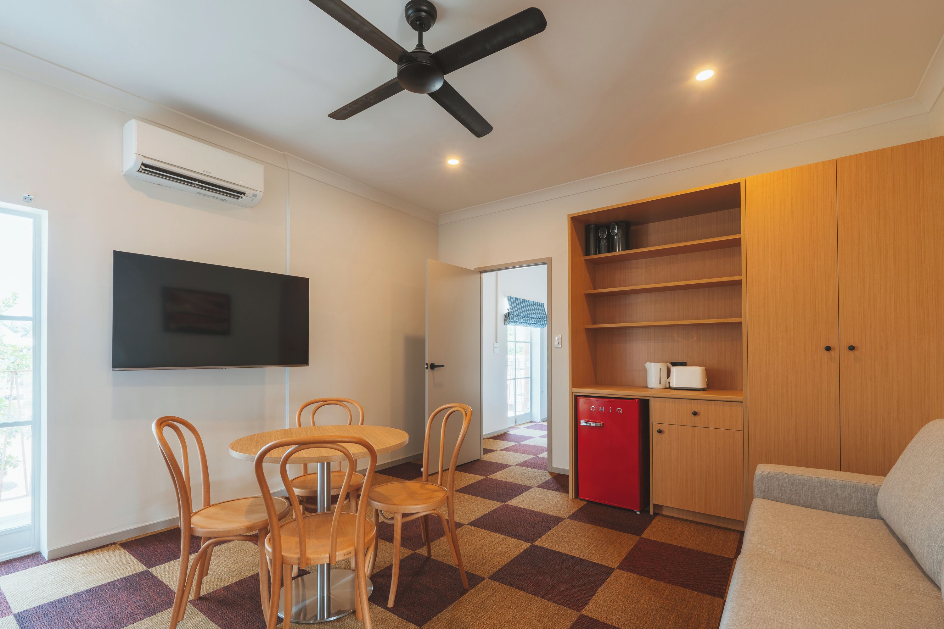 Une chambre moderne équipée d’une table à manger, de chaises, d’un mini-réfrigérateur rouge, d’une télévision et d’un ventilateur de plafond.