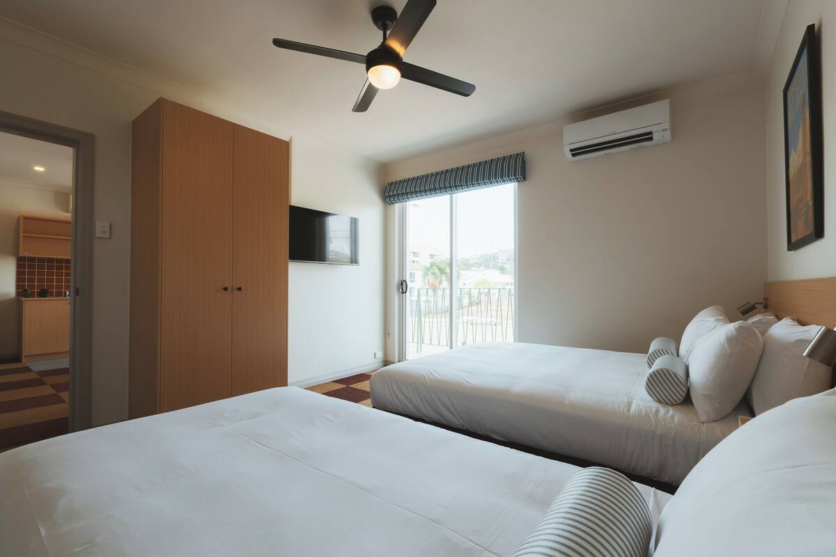 A hotel room with two beds, a ceiling fan, an air conditioning unit, a TV mounted on the wall, and a view of the outside through a sliding door.