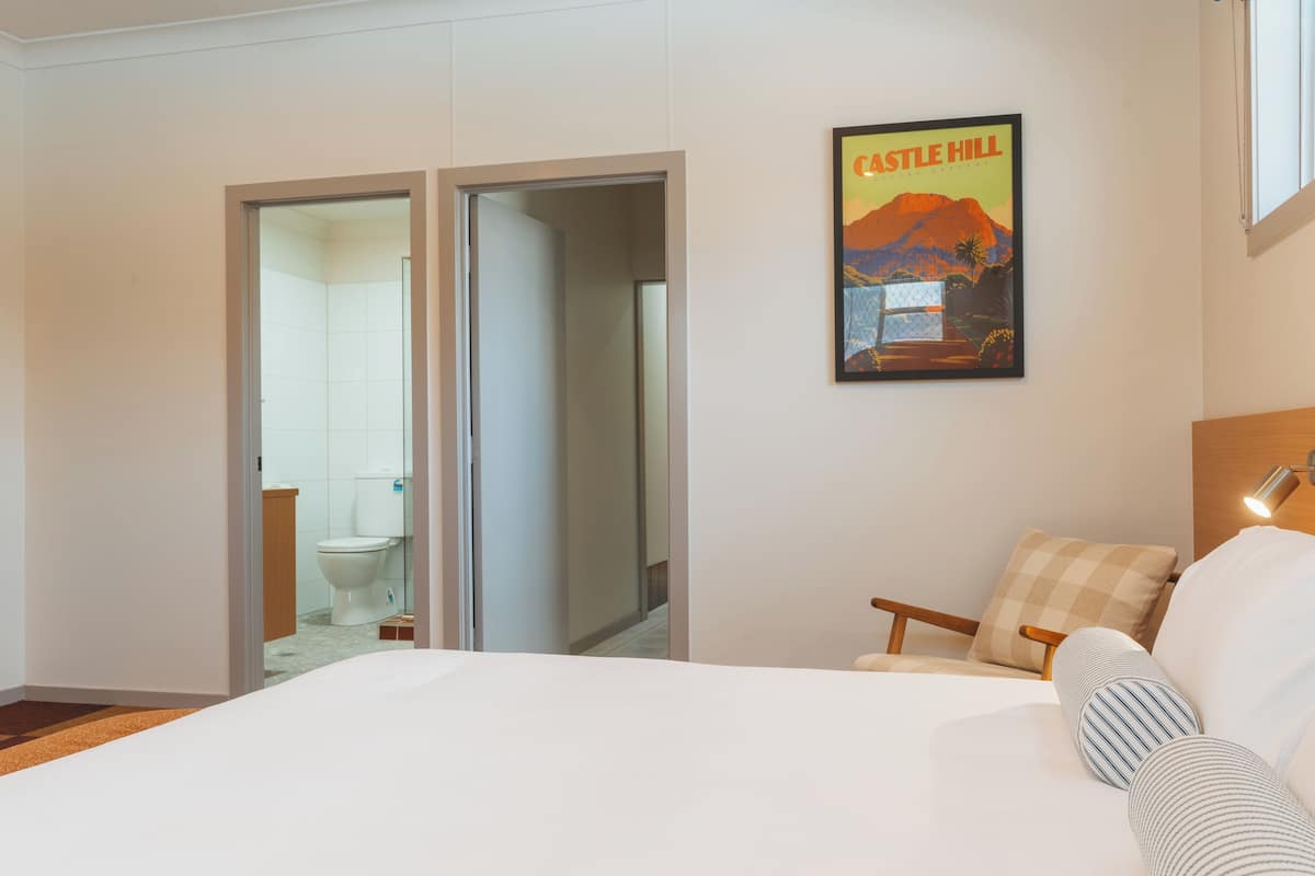A bedroom with a bed, a chair, a pillow, a poster on the wall, and a bathroom visible through an open door.