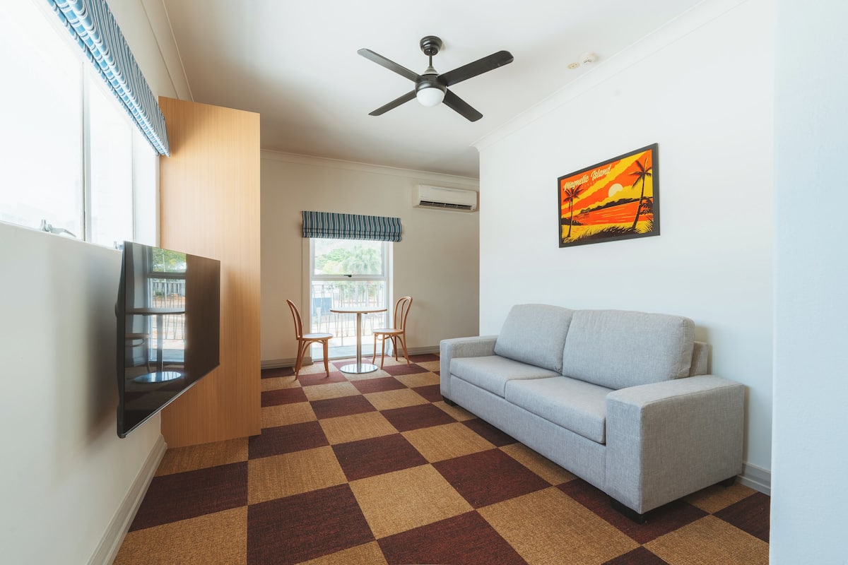 A living room with a sofa, a TV mounted on the wall, a ceiling fan, and a window with a view of the outdoors.