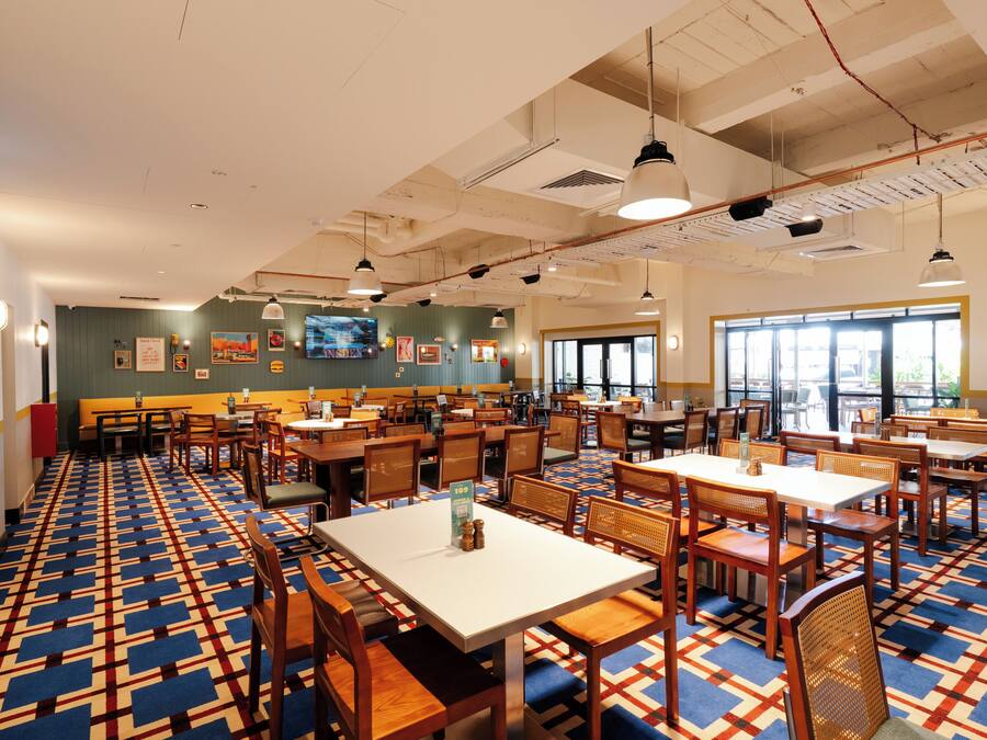 Un vaste espace dédié à la salle à manger, avec plusieurs tables et chaises, une moquette à motifs et une grande fenêtre laissant entrer la lumière naturelle.