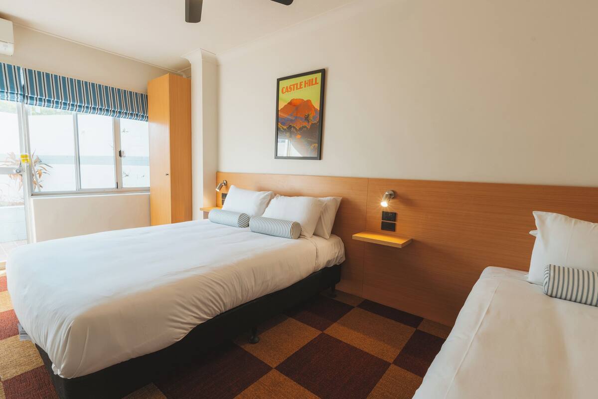 A hotel room with a large bed, a bedside table with a lamp, a poster on the wall, and a window with a view.