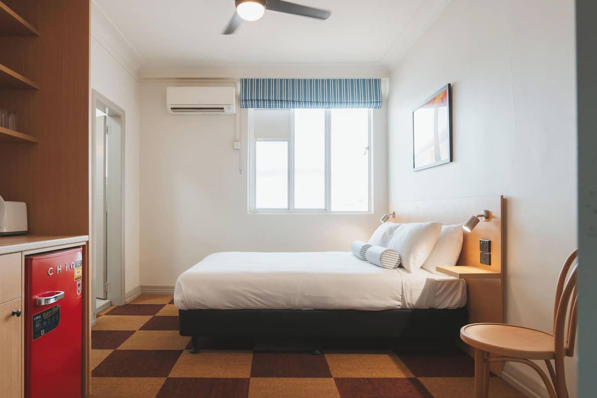A hotel room with a bed, a chair, a desk, a red minibar, a window with blinds, and an air conditioning unit.