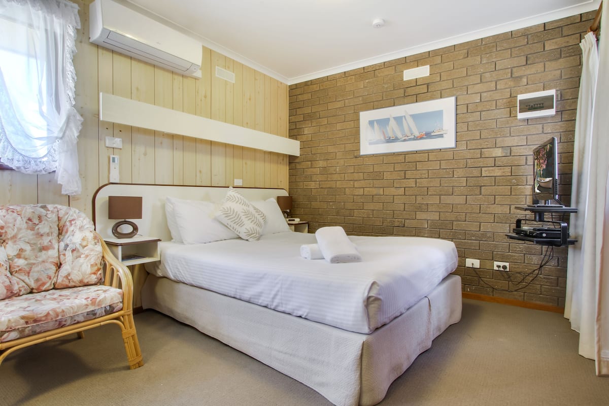 A room with a bed, a chair, a TV, and a brick wall.