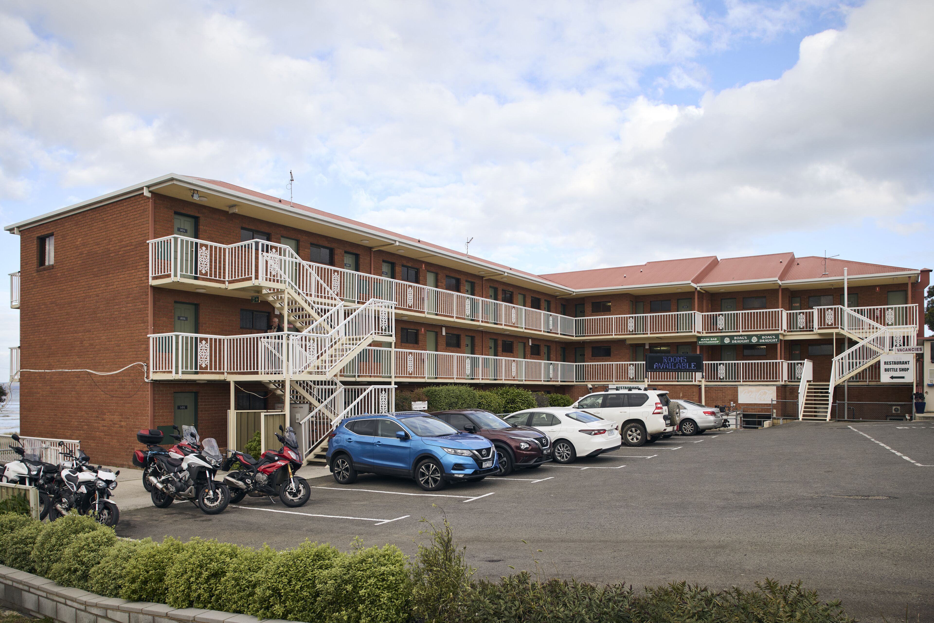 A multi-story hotel with a parking lot, a staircase to the upper floors, and a sign indicating rooms are available.