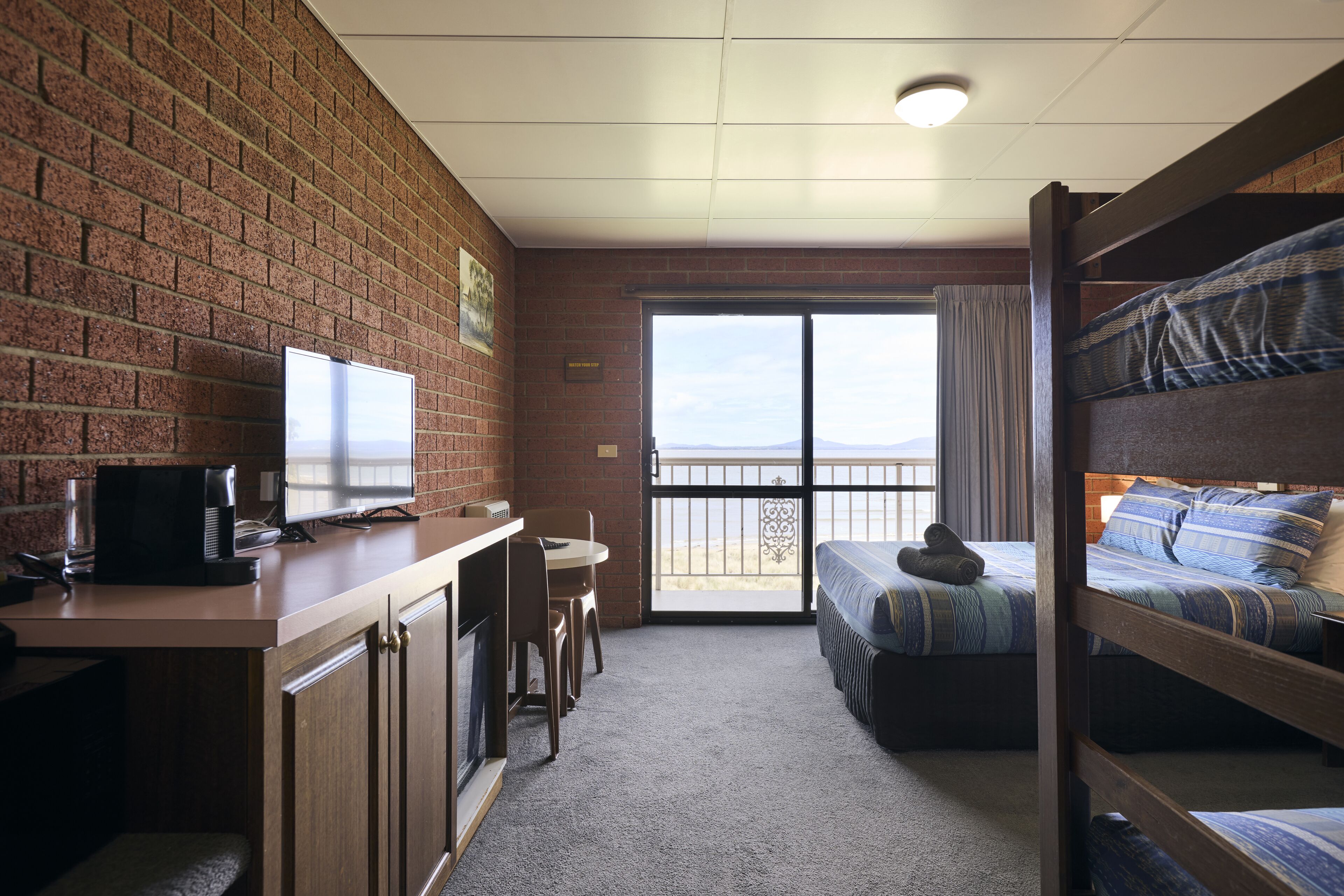 A room with a brick wall, a desk with a computer and a coffee maker, a bunk bed with blue bedding, and a large window with a view.