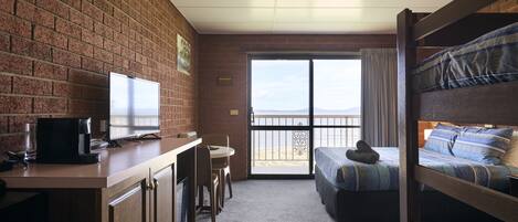 A room with a brick wall, a desk with a computer and a coffee maker, a bunk bed with blue bedding, and a large window with a view.