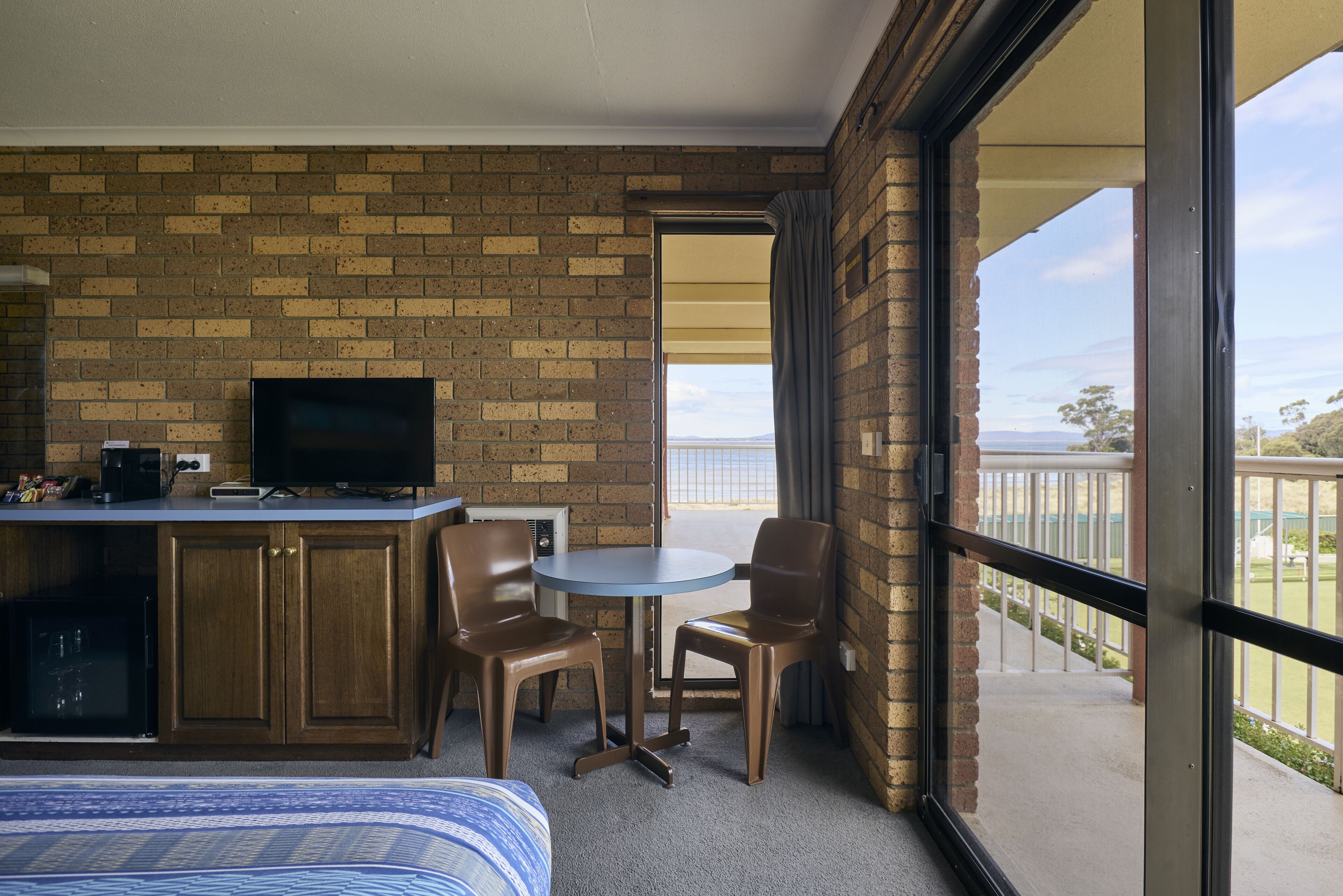 Habitación de hotel con pared de ladrillo, televisor, escritorio con silla, una mesita con dos sillas y balcón con vistas.